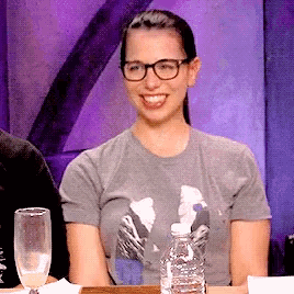 a woman wearing glasses sits at a table with a bottle of water and a glass of wine