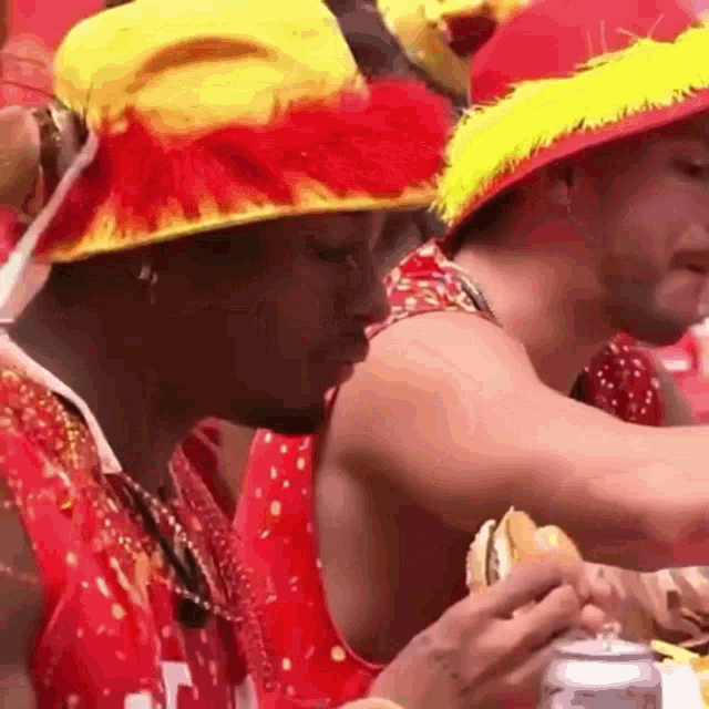 a man wearing a red and yellow hat is eating a hamburger and drinking a beer .