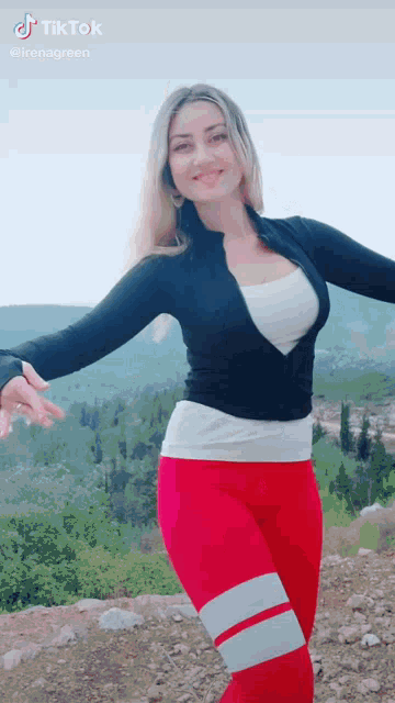 a woman in red leggings and a black jacket is standing on top of a rocky hill with her arms outstretched .