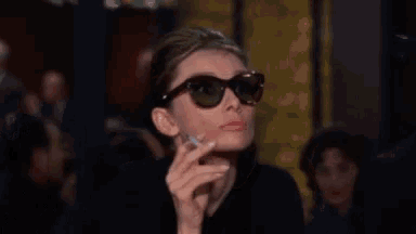 a woman wearing sunglasses is holding a cigarette in her hand and says gracious .