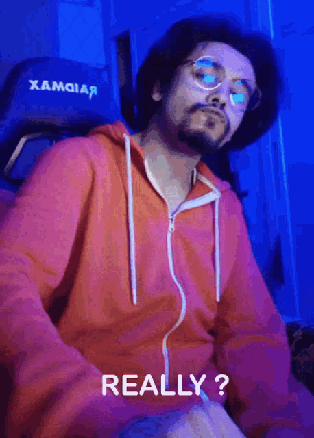 a man wearing glasses and a red hoodie is sitting in a gaming chair and says really