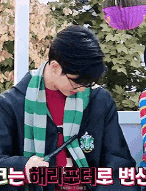 a young man wearing glasses and a green scarf is writing on a piece of paper with a pen .
