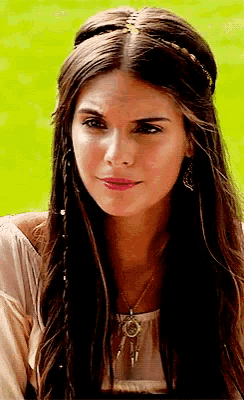a woman with braided hair wearing a necklace and earrings looks at the camera