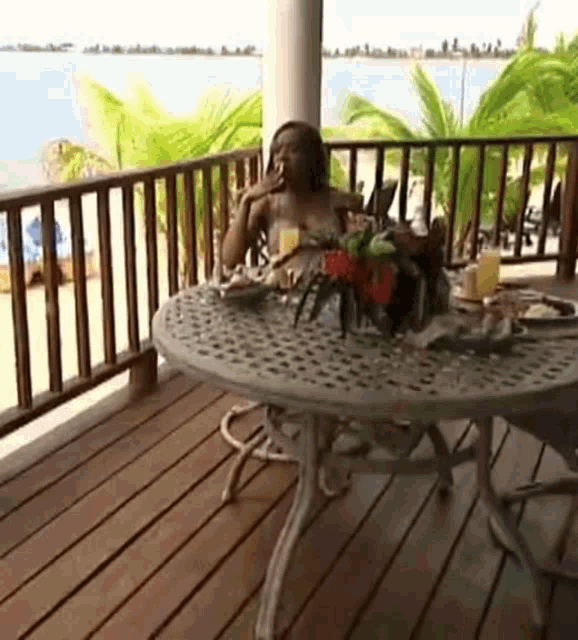 a woman is sitting at a table on a balcony drinking orange juice and smoking a cigarette