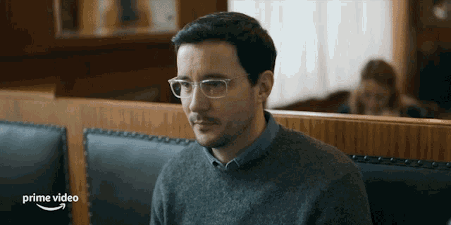 a man wearing glasses sits in a diner booth with a prime video logo behind him