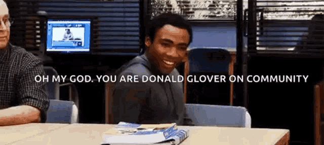 a man is sitting at a desk with a stack of books and says oh my god you are donald glover on community .
