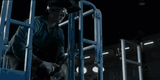 a man wearing a welding helmet and goggles is standing on a blue ladder .