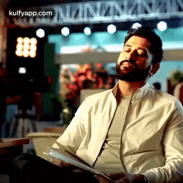 a man with a beard is sitting at a table reading a book and looking at his phone .