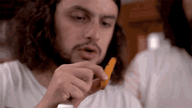 a man with long hair is eating a piece of carrot .
