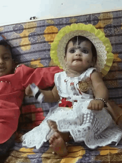 a baby girl wearing a white dress and a yellow hat sits next to a boy in a red shirt