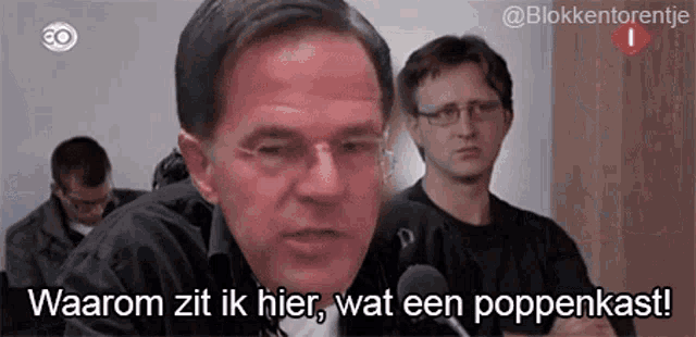 a man speaking into a microphone with the words waarom zit ik hier wat een poppenkast on the bottom