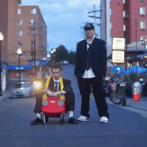 a man in a suit is pushing another man in a red toy car down a street