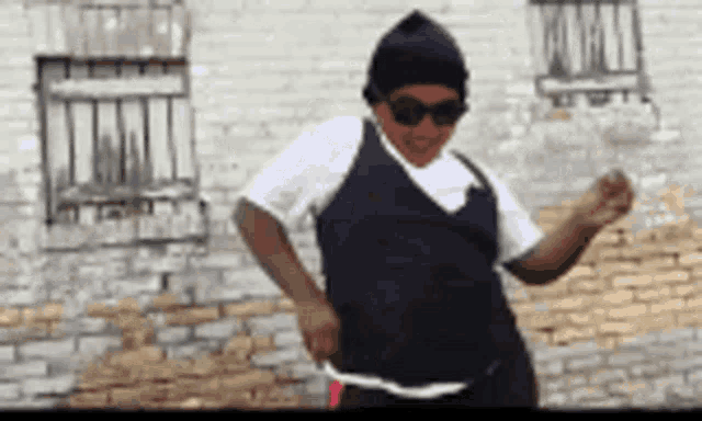a man wearing sunglasses and a beanie is dancing in front of a brick wall