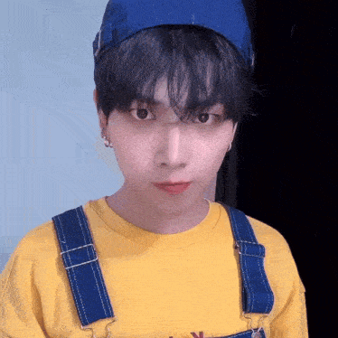 a young man wearing a yellow shirt and blue overalls is looking at the camera