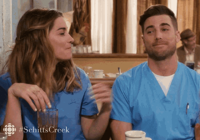 a man and a woman are sitting at a table with #schittscreek on the bottom