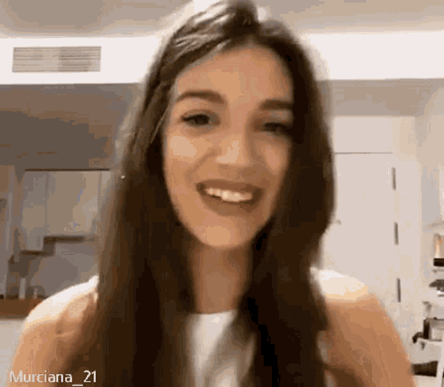a woman with long brown hair is smiling and looking at the camera in a kitchen .