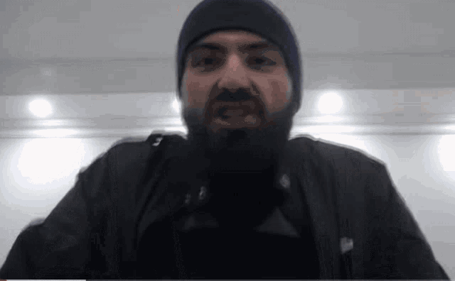 a man with a beard wearing a blue hat and a black jacket is looking at the camera .