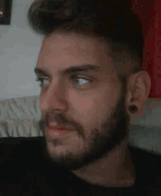 a man with a beard and ear plugs is sitting on a couch looking at the camera .