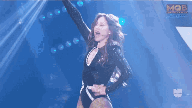 a woman in a black bodysuit stands on a stage with her arms in the air and a mob logo in the background