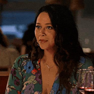 a woman wearing a floral shirt is sitting at a table with a glass of wine in front of her