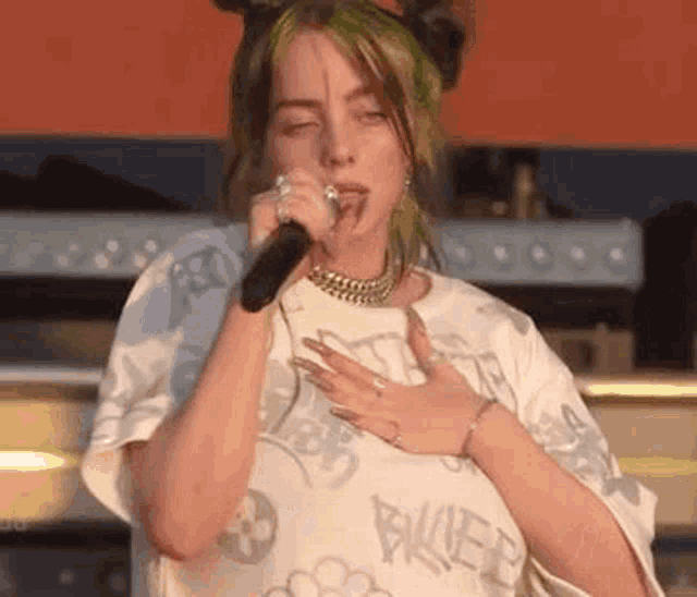 billie eilish is wearing a white t-shirt and holding a microphone in her hand .