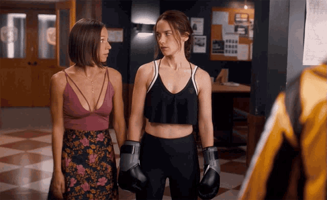 a woman wearing boxing gloves stands next to another woman in a gym