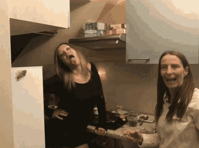 a woman sticking her tongue out while standing next to another woman in a kitchen