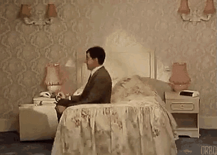 a man is sitting on a bed in a hotel room talking on a cell phone .