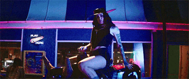 a woman in a cowboy hat is riding a motorcycle in front of a play here sign