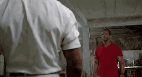 two men are standing next to each other in a room . one is wearing a red shirt .
