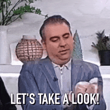 a man in a suit is saying `` let 's take a look '' while holding something in his hand .