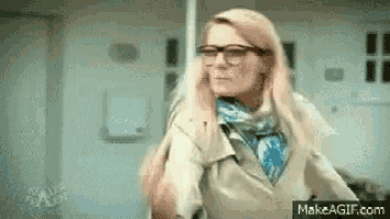 a woman wearing glasses , a scarf and a trench coat is standing in a room .