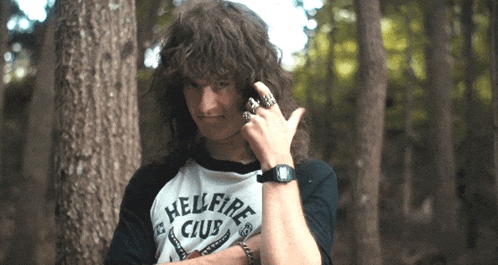 a man wearing a shirt that says hellfire club is standing in the woods