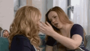 two women are sitting on a couch talking to each other and one is touching the other 's face .