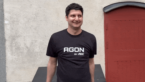 a man wearing a t-shirt that says agon by aoc stands in front of a red door