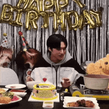 a man is sitting at a table with a birthday cake and balloons that say happy birthday behind him