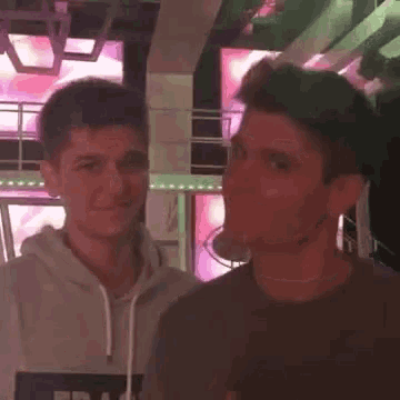 two young men are standing next to each other in a room with a pink light behind them .