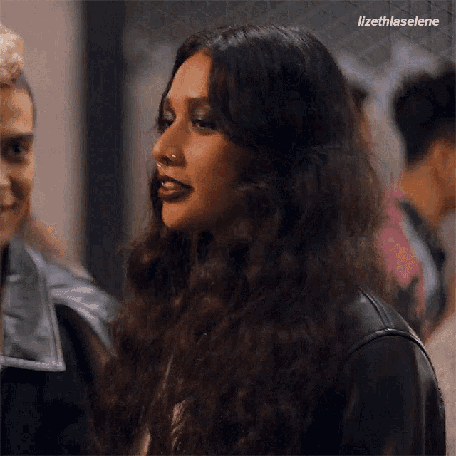 a woman with long curly hair and a nose ring is wearing a leather jacket and talking to a man .