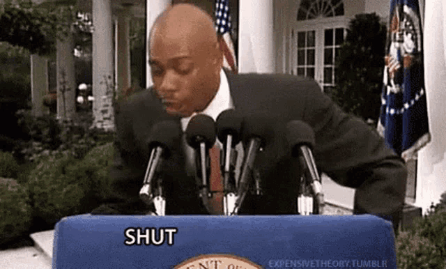a man in a suit and tie is standing behind a podium with microphones and a sign that says shut .
