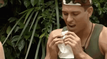 a man is eating a sandwich in the jungle while wearing a hat .