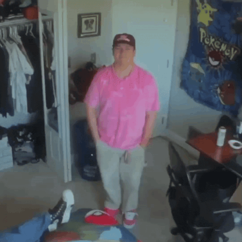a man in a pink shirt stands in a room with a pokemon tapestry on the wall