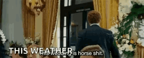 a man in a suit is standing in a room with flowers and a sign that says `` this weather is horse shit . ''