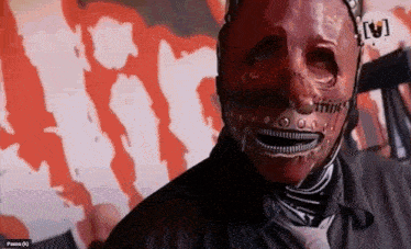 a man wearing a red mask with a skull on it looks at the camera with a pause button in the corner