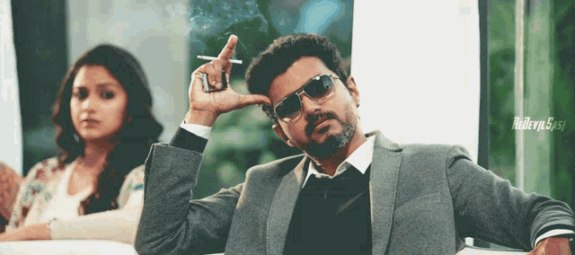 a man in a suit and sunglasses smoking a cigarette with a woman in the background