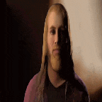 a man with long hair and a beard is standing in a dark room looking at the camera .
