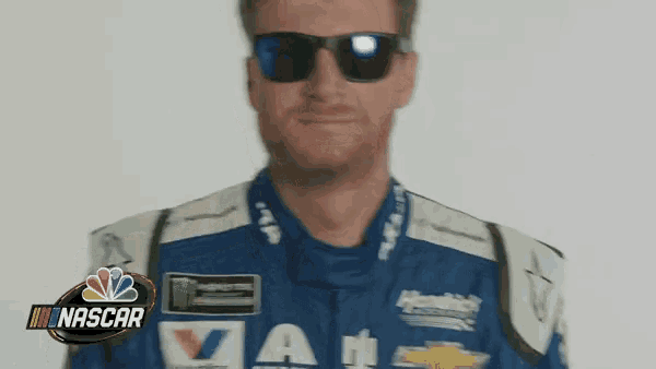 a man wearing sunglasses and a nascar uniform is standing in front of a white background .