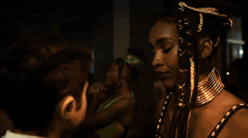 a woman with braids and a gold necklace talks to a man in a dark room