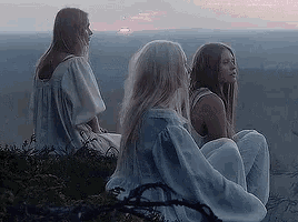 a group of three women are sitting on top of a hill looking at the sunset .