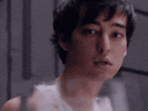 a young man in a white tank top is looking at the camera with a serious look on his face .