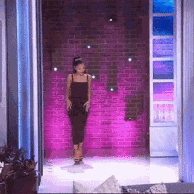 a woman in a black dress is standing in front of a brick wall with purple lights behind her .